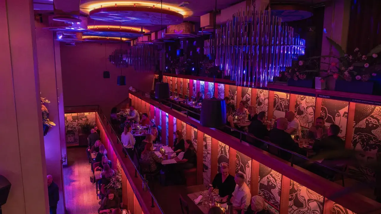 En livlig restaurang med två våningar, upplyst av lila och orange ljus. Gäster sitter vid borden och njuter av kvällen. Biblioteket live!