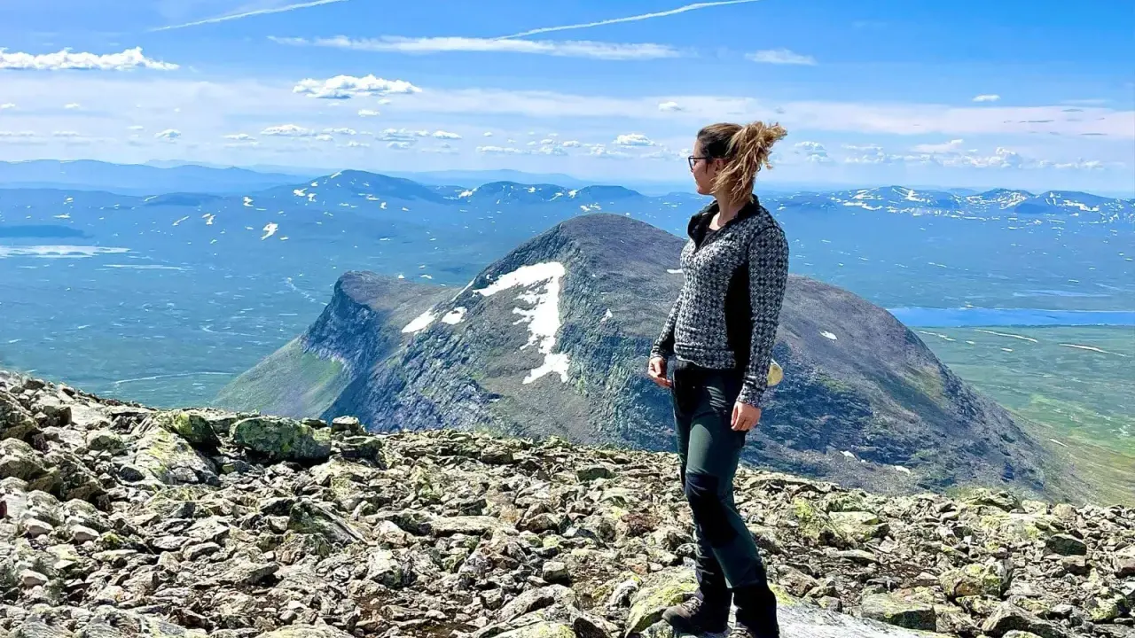 En kvinna njuter av utsikten från en stenig topp efter en helags topptur. Vackert landskap med snöklädda berg och blå himmel.