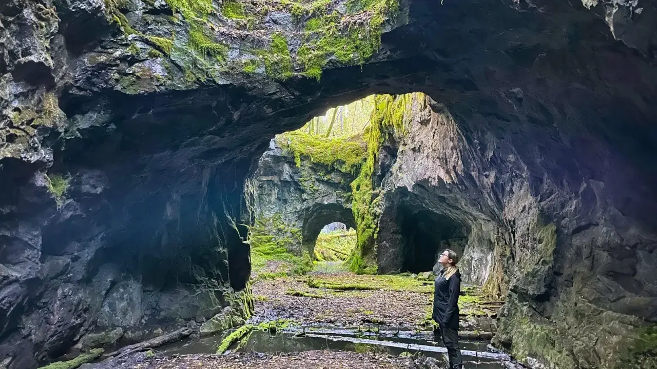 En kvinna står i en mossig grotta, en del av **Tilas stoll**, och blickar upp mot ljuset som sipprar in.