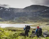 Två vandrare med ryggsäckar går längs en sjö i ett bergigt landskap. En del av snö syns på topparna. Perfekt för vandringsleder Sverige.