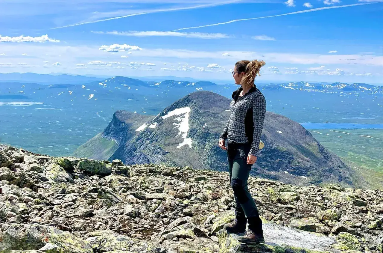 En kvinna njuter av utsikten från en stenig topp efter en helags topptur. Vackert landskap med snöklädda berg och blå himmel.