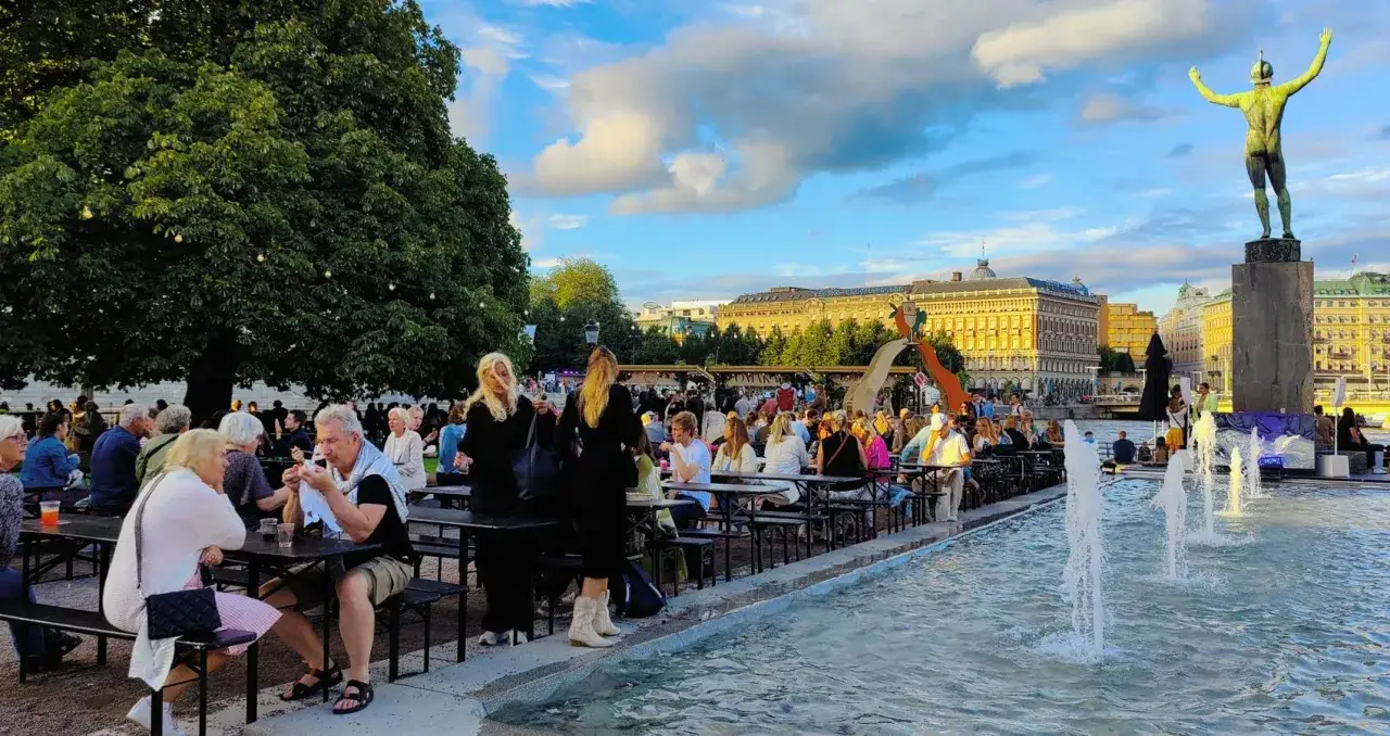 Folk njuter av mat och dryck vid bord utomhus, med en fontän och en staty i bakgrunden. En livlig atmosfär som påminner om kulturfestivalen Stockholm.