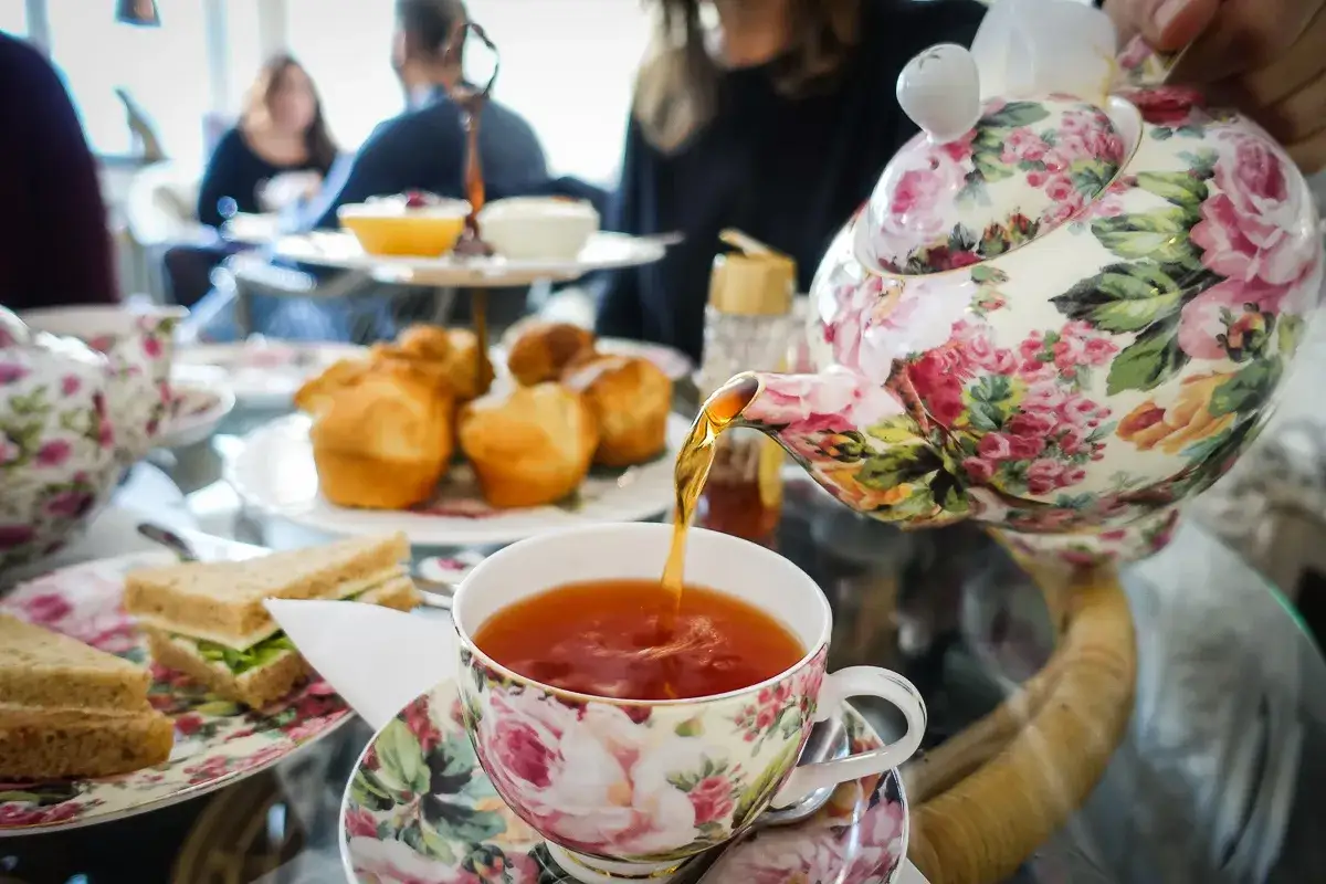 En blommig tekanna häller upp te i en matchande kopp. Smörgåsar och scones syns på bordet, redo för ett härligt afternoon tea.