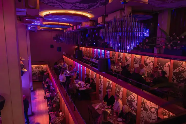 En livlig restaurang med två våningar, upplyst av lila och orange ljus. Gäster sitter vid borden och njuter av kvällen. Biblioteket live!